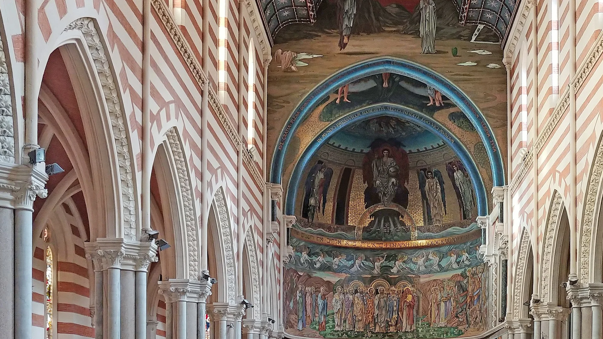 Der Blick geht ein römisches Kirchenschiff entlang, das in Rot und Creme gebändert ist, unter einer blauen, mit Sternen übersäten Decke, bis zur Apsis, die in goldenen Mosaiken erstrahlt.