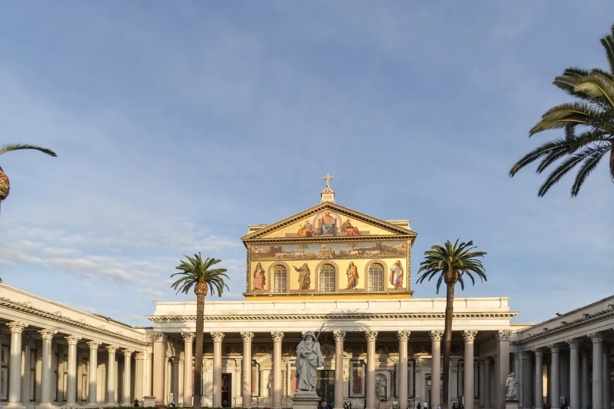 Ein Hof mit weißen Säulen und Palmen, der zu einer Basilikafassade mit einem von Goldmosaik gekrönten Giebel führt.